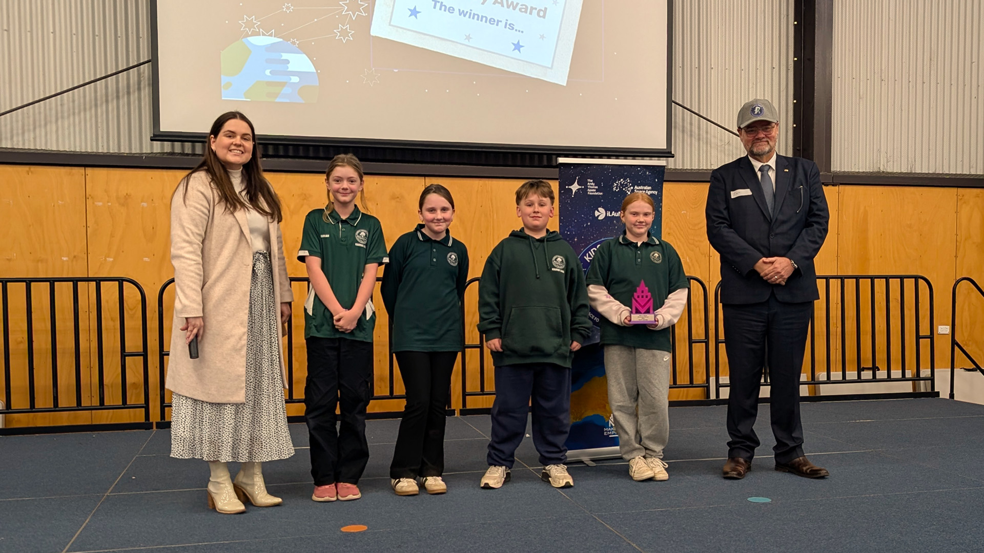 Two guest judges and four school kids from Riverdale Primary School standing on stage accepting an award for the 2025 Kids in Space program final.