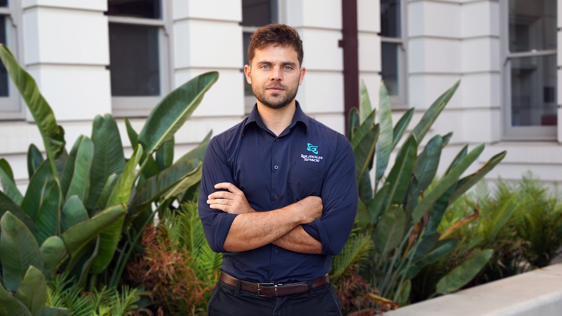 Male standing in front of green shrubs with arms crossed.