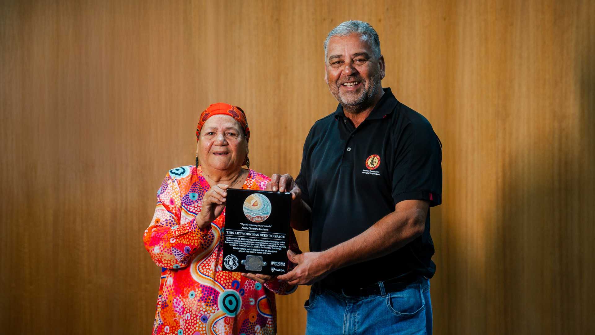 From Orbit to Country Two people standing indoors holding a commemorative Indigenous artwork plaque together.