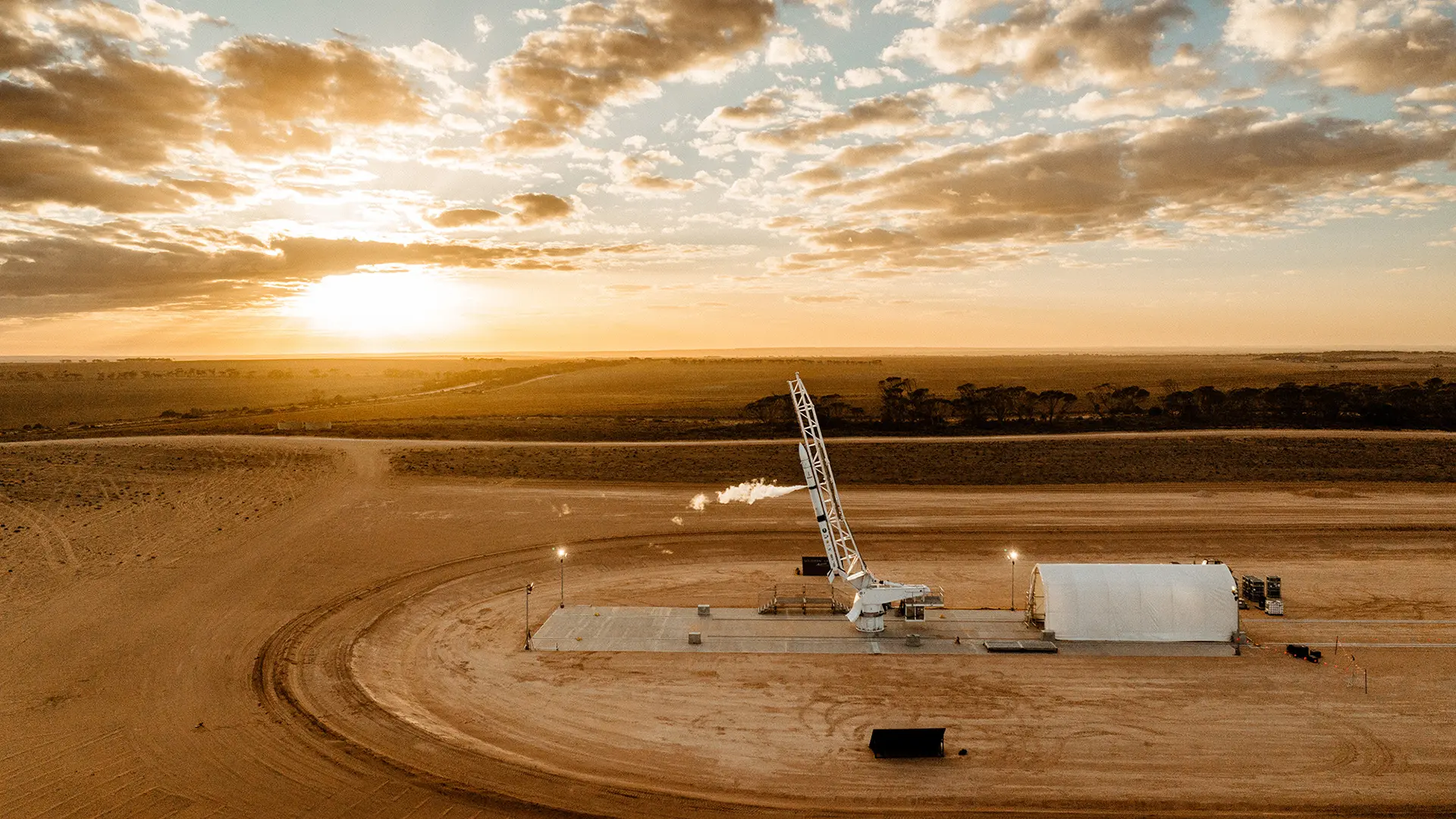 A rocket positioned on a launch pad in an open desert landscape at sunset, with soft clouds across the sky and a large white shelter beside the launch area