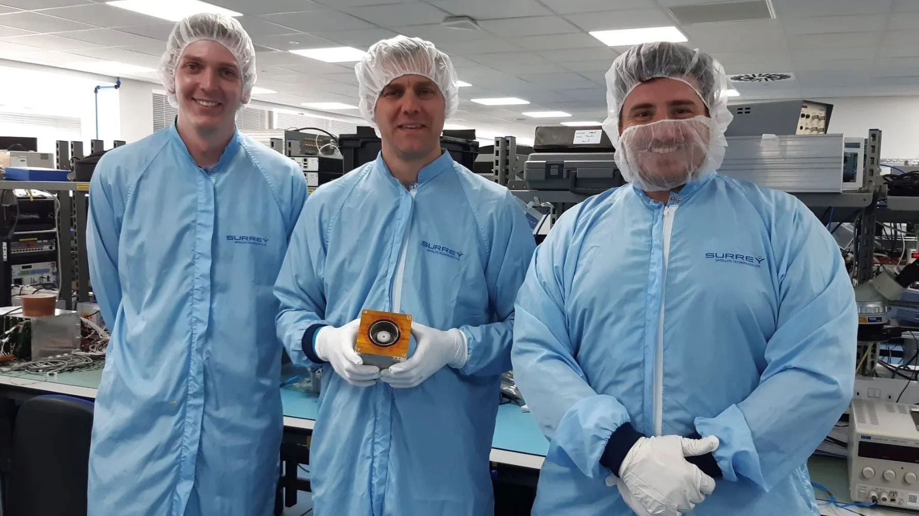 Three men in blue lab coats with hair nets on holding a satellite propulsion component