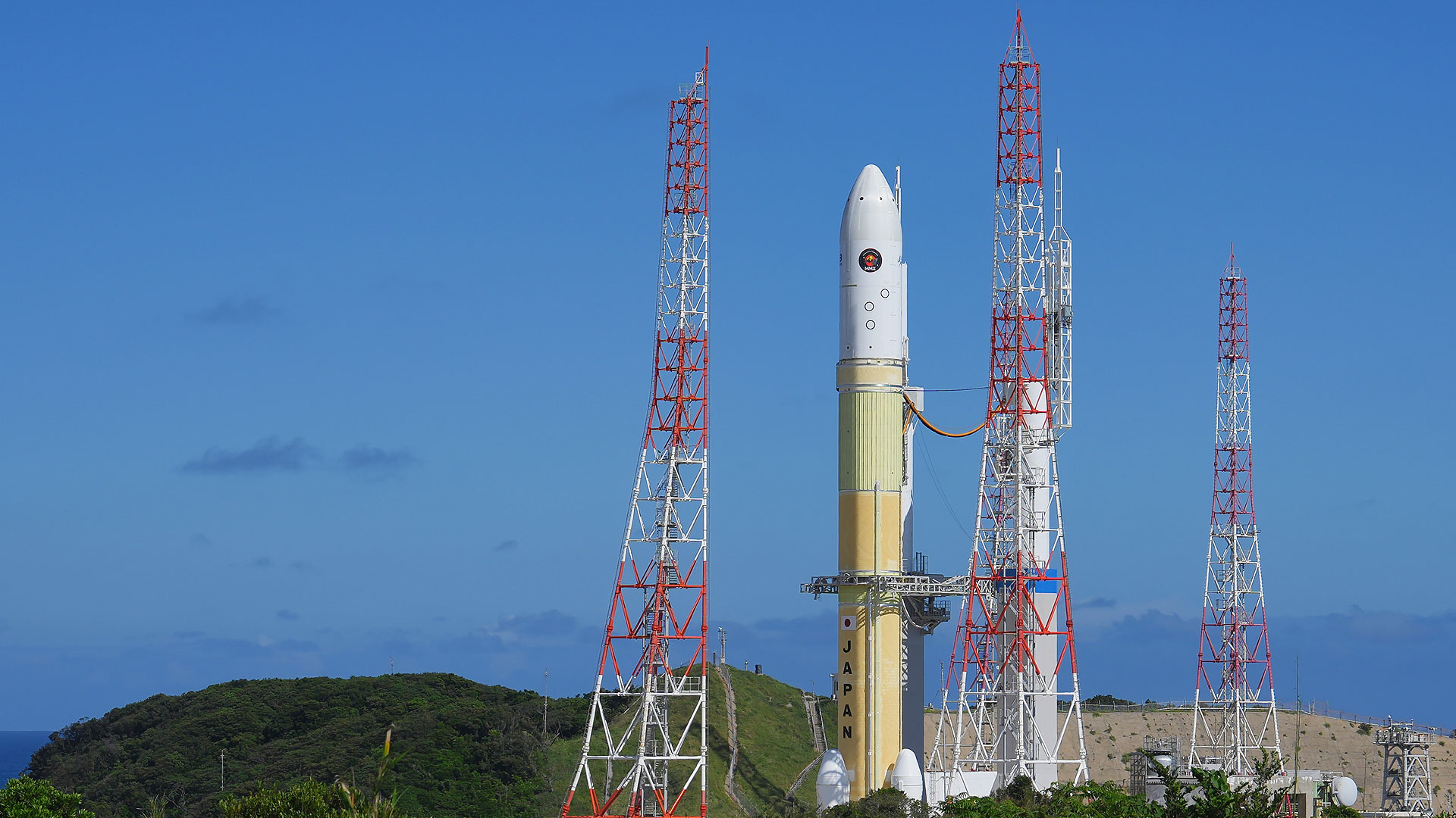 Jaxa-photo Japan Aerospace Exploration Agency (JAXA) MMX mission rocket stands on a launch pad surrounded by red and white service towers before launch, against a clear blue sky.