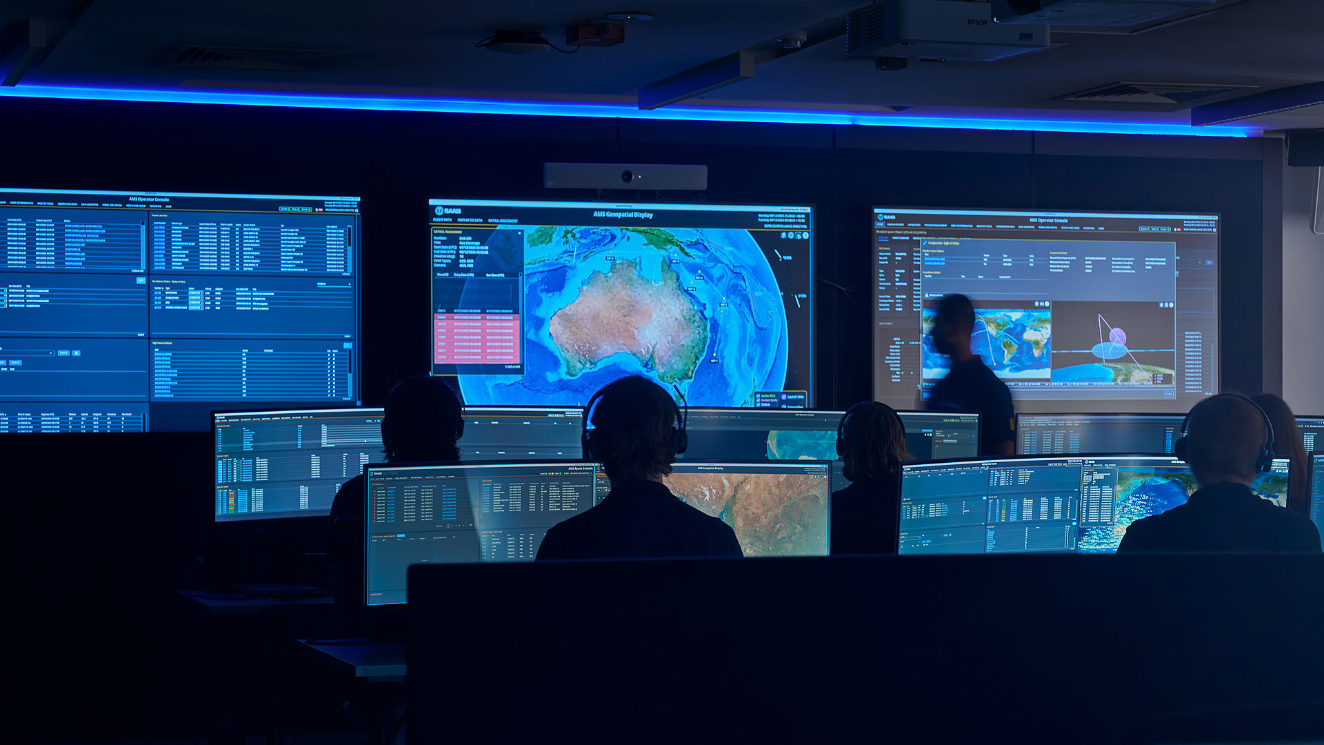 Personnel monitor satellite and mission data on multiple screens in an operations centre displaying maps, telemetry and space systems information.